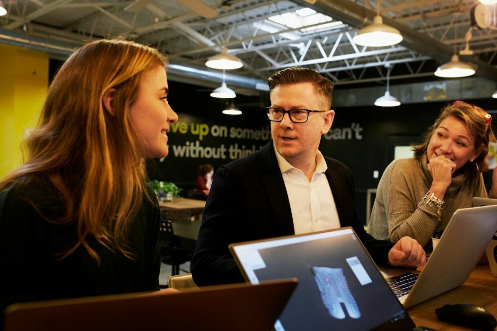 A man in glasses and a dark blazer talks with two smiling women across a table of open laptops in a busy industrial-style office.