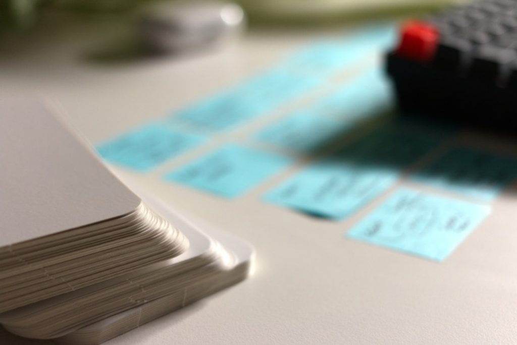 A stack of blank white index cards in the foreground beside rows of small blue sticky notes with handwritten labels sorted into columns on a white desk, with the corner of a black keyboard blurred in the background.