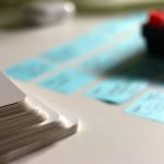 A stack of blank white index cards in the foreground beside rows of small blue sticky notes with handwritten labels sorted into columns on a white desk, with the corner of a black keyboard blurred in the background.
