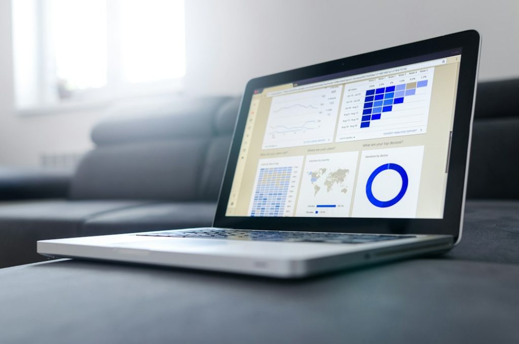An open silver laptop on a dark grey couch, its screen showing a web analytics dashboard with line charts, a blue cohort table, a world map and a donut chart.