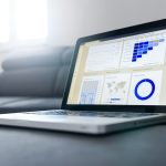 An open silver laptop on a dark grey couch, its screen showing a web analytics dashboard with line charts, a blue cohort table, a world map and a donut chart.