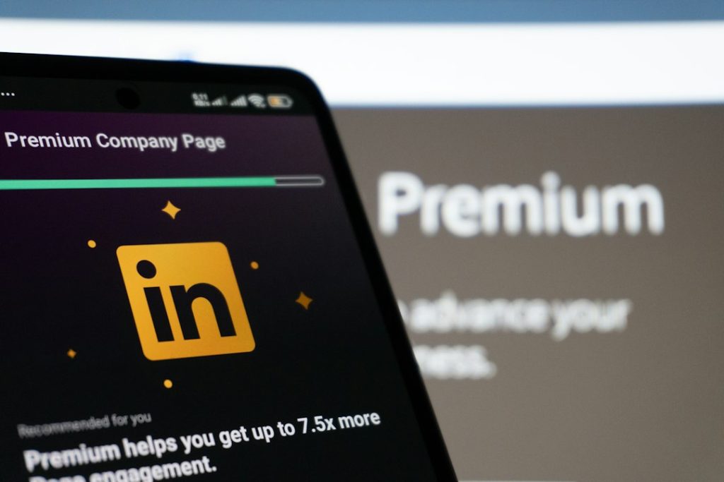 A smartphone angled in front of a blurred monitor, its dark screen showing a LinkedIn Premium Company Page prompt with the gold LinkedIn logo.