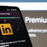 A smartphone angled in front of a blurred monitor, its dark screen showing a LinkedIn Premium Company Page prompt with the gold LinkedIn logo.