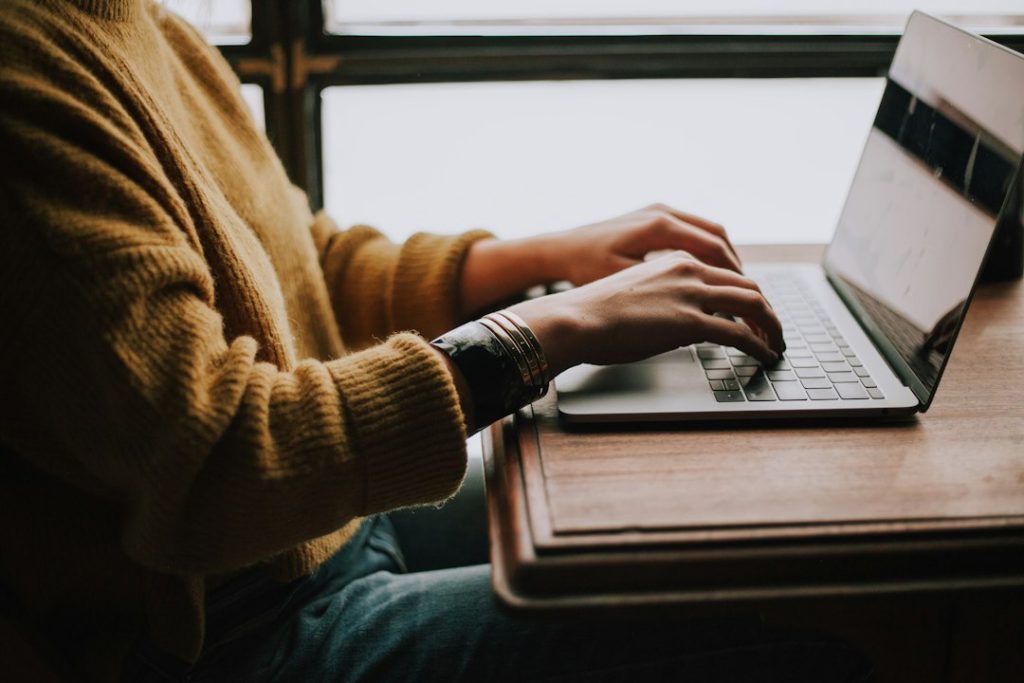 A person in a mustard knit sweater typing on a silver laptop resting on a wooden desk beside a window.