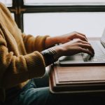 A person in a mustard knit sweater typing on a silver laptop resting on a wooden desk beside a window.
