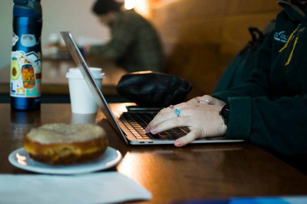 Close-up of hands with rings typing on a laptop at a wooden cafe table, next to a takeaway coffee cup, a sticker-covered water bottle, and a donut on a plate.