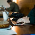 Close-up of hands with rings typing on a laptop at a wooden cafe table, next to a takeaway coffee cup, a sticker-covered water bottle, and a donut on a plate.