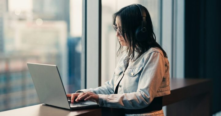 A woman in a denim jacket and headphones types on a laptop at a standing desk beside a tall window with a blurred city skyline behind her.