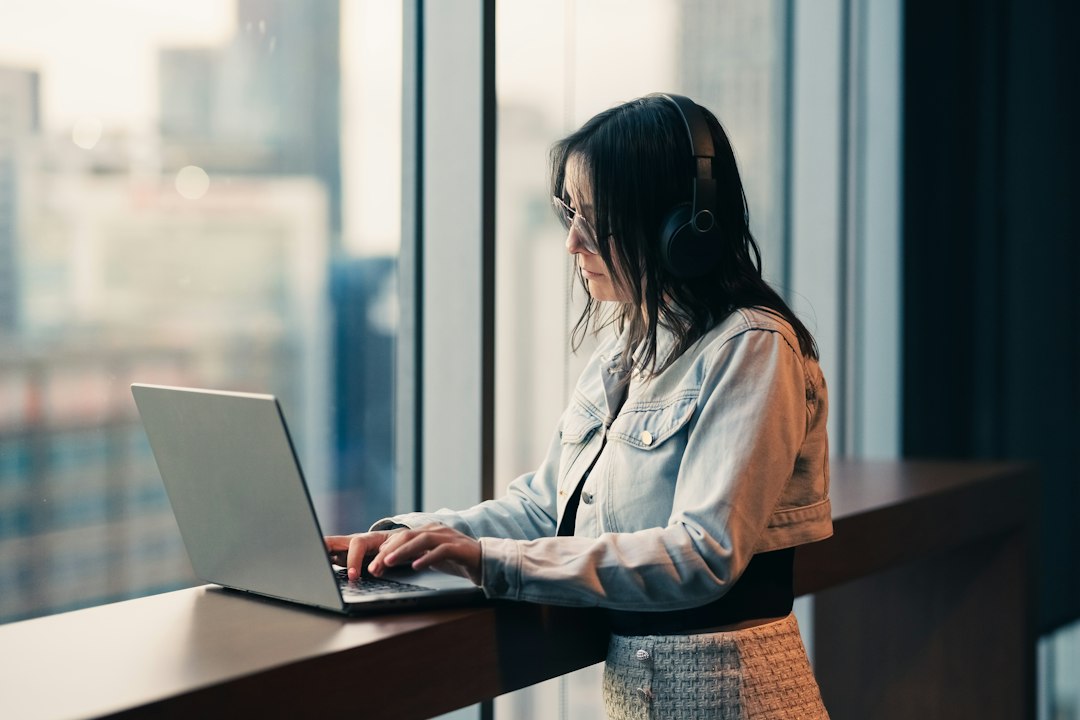 A woman in a denim jacket and headphones types on a laptop at a standing desk beside a tall window with a blurred city skyline behind her.