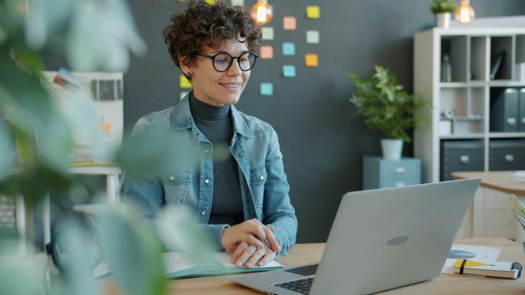 A smiling woman with curly hair and round glasses sits at a wooden desk in a bright office, looking at an open laptop with a notebook in front of her and colored sticky notes on the wall behind.