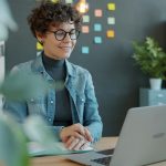 A smiling woman with curly hair and round glasses sits at a wooden desk in a bright office, looking at an open laptop with a notebook in front of her and colored sticky notes on the wall behind.