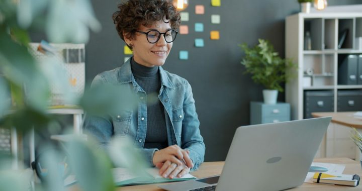 A smiling woman with curly hair and round glasses sits at a wooden desk in a bright office, looking at an open laptop with a notebook in front of her and colored sticky notes on the wall behind.