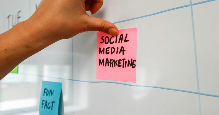 A hand pressing a pink sticky note reading "Social Media Marketing" onto a whiteboard planning grid, with a blue "Fun Fact" note below it.