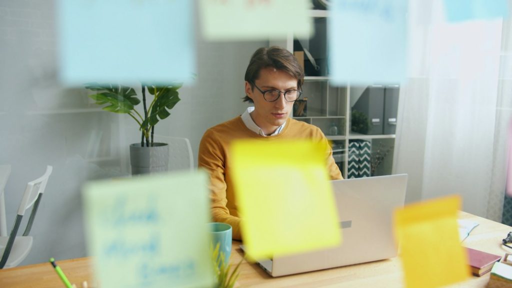 A man in glasses and a mustard sweater works on a laptop at a wooden desk, seen through a glass wall covered in blurred colorful sticky notes.