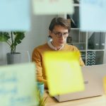 A man in glasses and a mustard sweater works on a laptop at a wooden desk, seen through a glass wall covered in blurred colorful sticky notes.