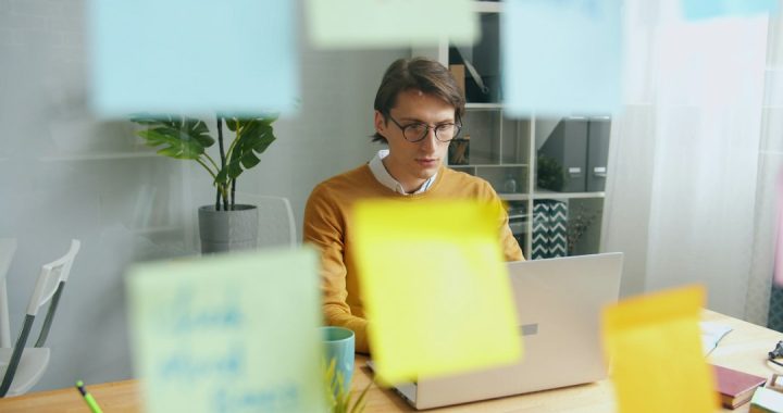 A man in glasses and a mustard sweater works on a laptop at a wooden desk, seen through a glass wall covered in blurred colorful sticky notes.