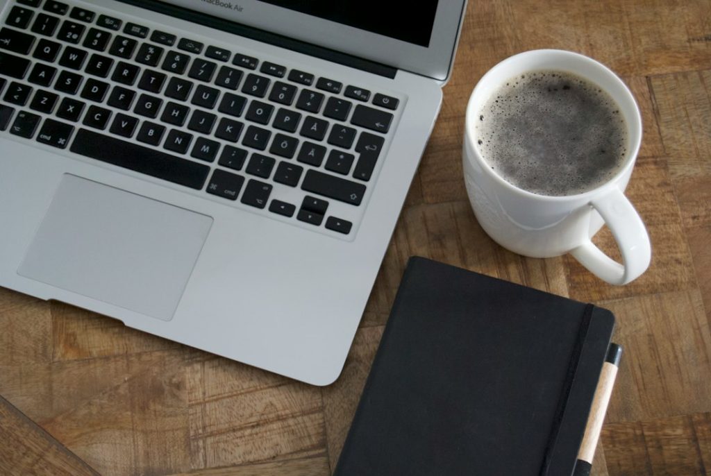 Overhead view of an open silver laptop keyboard beside a white mug of coffee and a closed black notebook with a pen, on a wooden table.