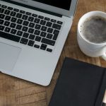 Overhead view of an open silver laptop keyboard beside a white mug of coffee and a closed black notebook with a pen, on a wooden table.