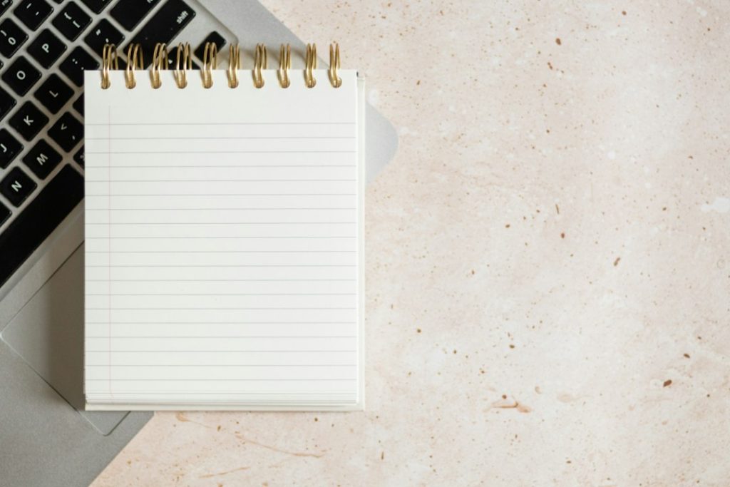 Blank spiral-bound lined notepad resting on a silver laptop keyboard, on a beige speckled stone surface.