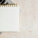 Blank spiral-bound lined notepad resting on a silver laptop keyboard, on a beige speckled stone surface.