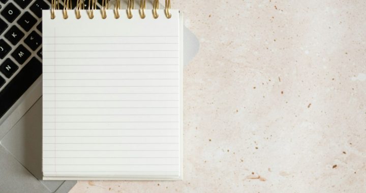 Blank spiral-bound lined notepad resting on a silver laptop keyboard, on a beige speckled stone surface.