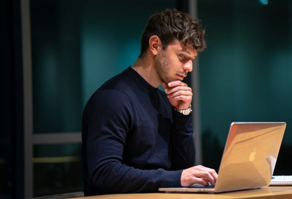 A man in a dark sweater rests his chin on his hand and studies a laptop screen at a desk in a dim room.