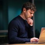 A man in a dark sweater rests his chin on his hand and studies a laptop screen at a desk in a dim room.