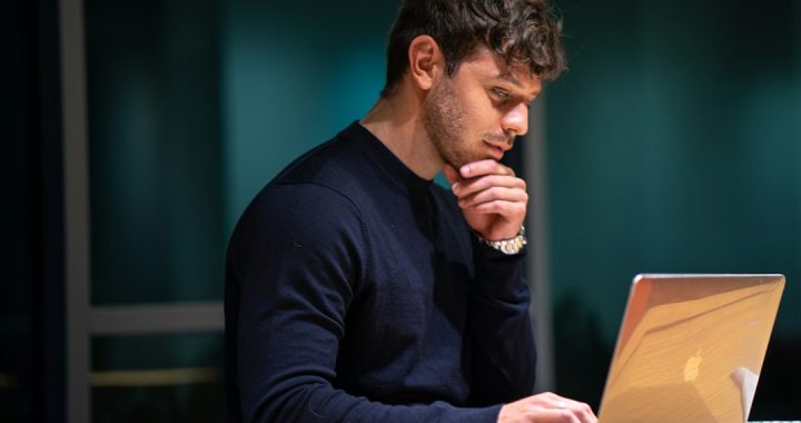 A man in a dark sweater rests his chin on his hand and studies a laptop screen at a desk in a dim room.