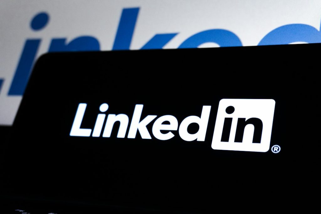 White LinkedIn logo on a black screen, with a larger blurred blue LinkedIn wordmark in the background.