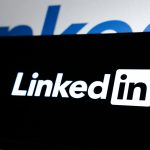 White LinkedIn logo on a black screen, with a larger blurred blue LinkedIn wordmark in the background.
