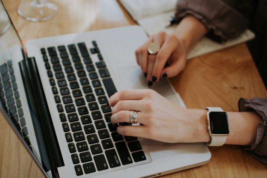 Close-up of a person's hands typing on a silver laptop keyboard at a wooden desk, with a smartwatch on one wrist.