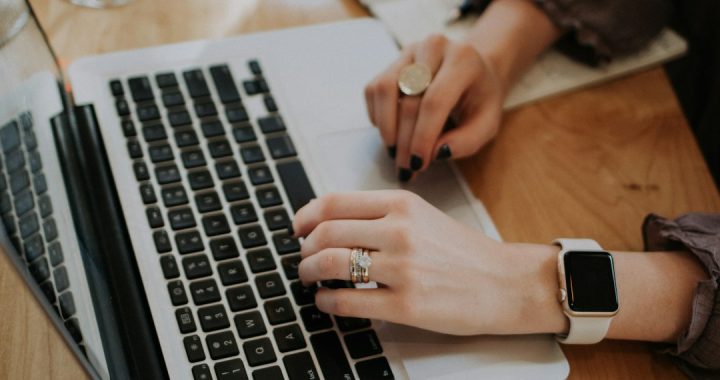 Close-up of a person's hands typing on a silver laptop keyboard at a wooden desk, with a smartwatch on one wrist.