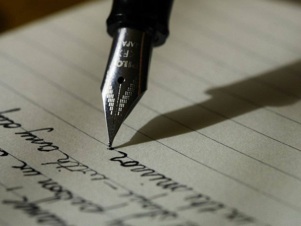 Close-up of a fountain pen nib writing cursive handwriting on lined paper, with the pen's shadow falling across the page.
