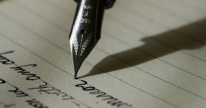 Close-up of a fountain pen nib writing cursive handwriting on lined paper, with the pen's shadow falling across the page.