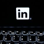 The white LinkedIn logo on a black laptop screen above a backlit keyboard in a dark room.