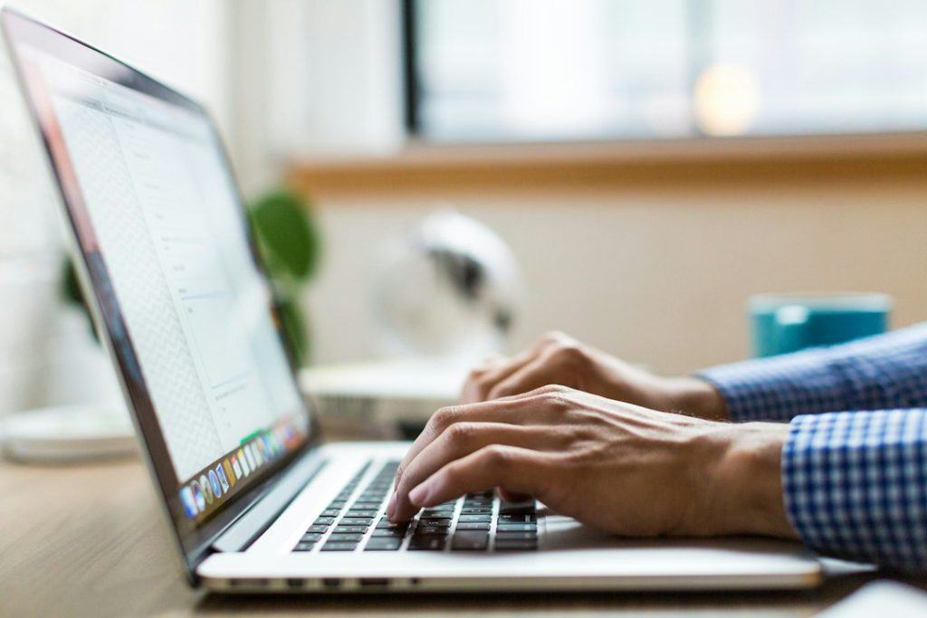 Close-up of a man's hands in a blue gingham shirt typing on a silver laptop at a wooden desk, with a window and a teal mug blurred behind.