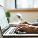 Close-up of a man's hands in a blue gingham shirt typing on a silver laptop at a wooden desk, with a window and a teal mug blurred behind.