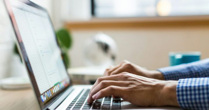 Close-up of a man's hands in a blue gingham shirt typing on a silver laptop at a wooden desk, with a window and a teal mug blurred behind.