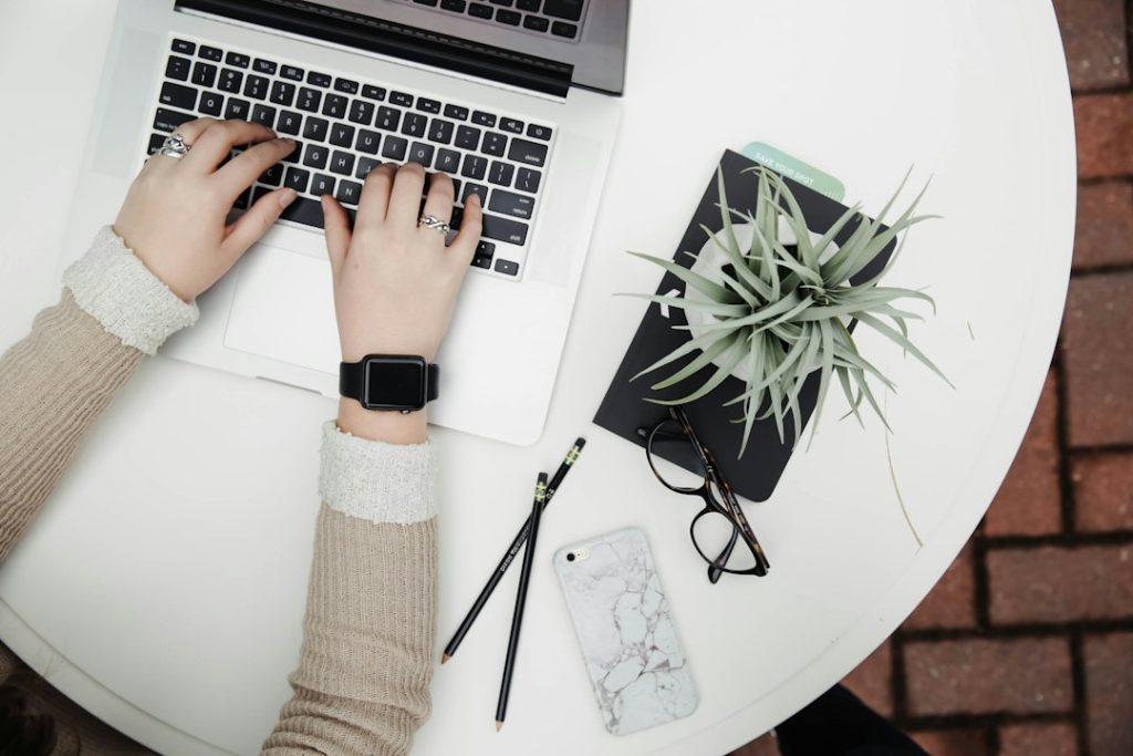 Overhead view of two hands typing on a silver laptop at a white round table, with a black notebook, an air plant, glasses, two pencils and a marble phone case beside it.