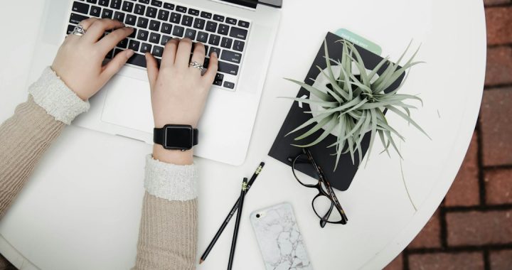 Overhead view of two hands typing on a silver laptop at a white round table, with a black notebook, an air plant, glasses, two pencils and a marble phone case beside it.