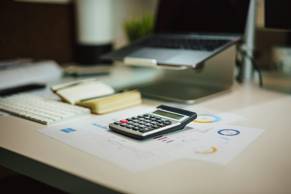 A black calculator resting on printed charts and graphs on a desk, with a notebook, white keyboard and laptop on a stand out of focus behind it.