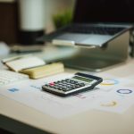 A black calculator resting on printed charts and graphs on a desk, with a notebook, white keyboard and laptop on a stand out of focus behind it.