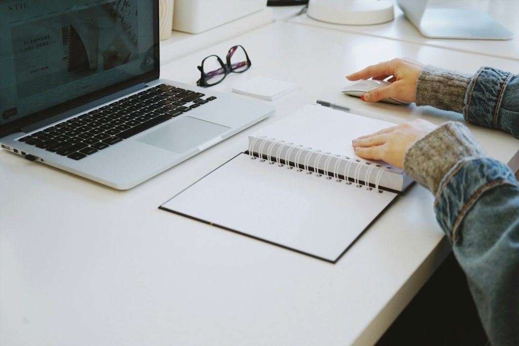 A person at a white desk with a laptop, an open spiral notebook, and a pair of glasses, one hand on a mouse and the other resting on the notebook.