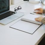A person at a white desk with a laptop, an open spiral notebook, and a pair of glasses, one hand on a mouse and the other resting on the notebook.