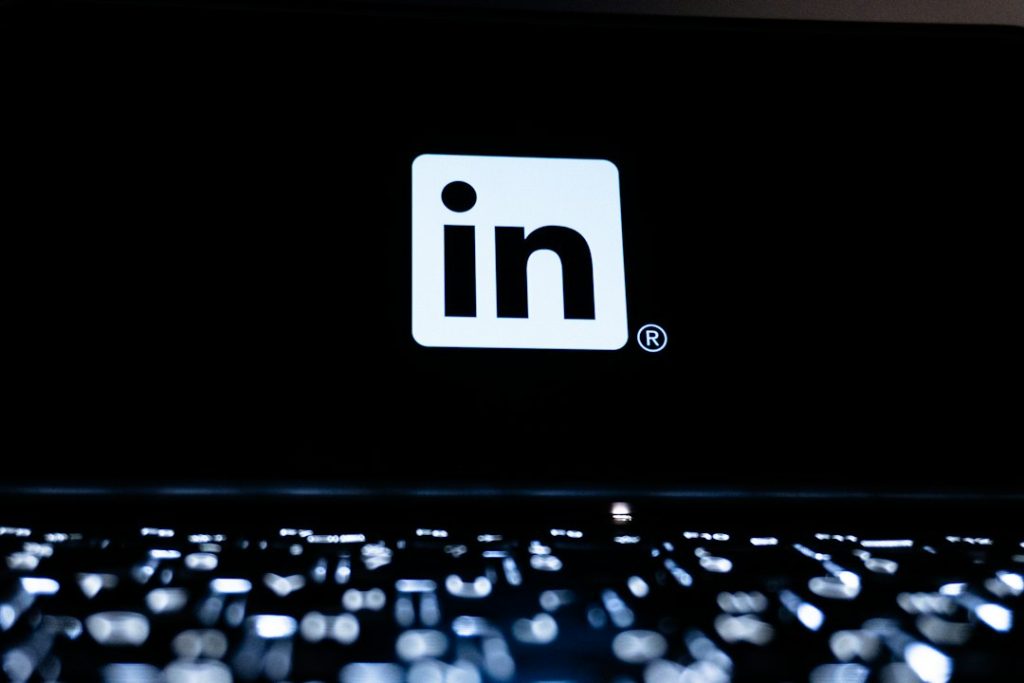 White LinkedIn logo on a black laptop screen, with a backlit keyboard glowing out of focus in the foreground.