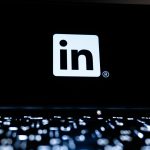 White LinkedIn logo on a black laptop screen, with a backlit keyboard glowing out of focus in the foreground.