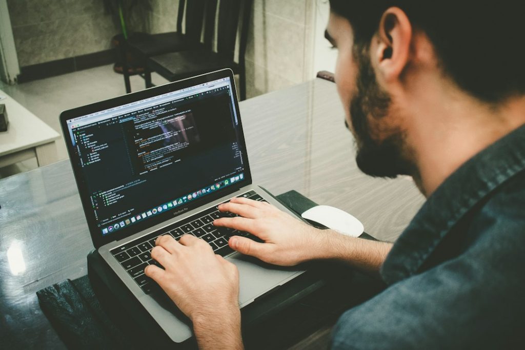 A bearded man in a dark denim shirt types on a MacBook Pro with a code editor open, seen over his shoulder at a wooden desk.