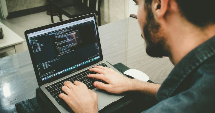 A bearded man in a dark denim shirt types on a MacBook Pro with a code editor open, seen over his shoulder at a wooden desk.