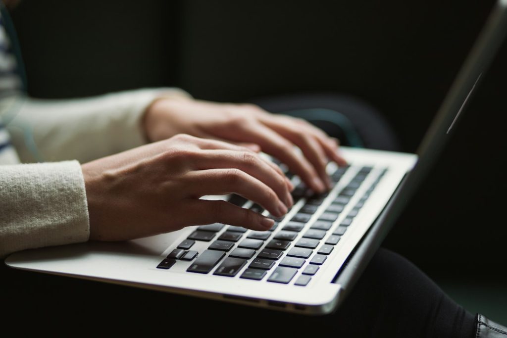 Close-up of a person in a cream knit sweater typing on a silver laptop resting on their lap, against a dark background.