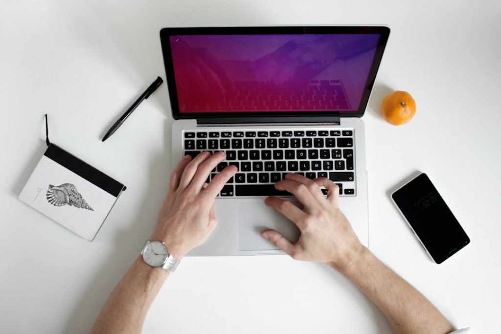 Overhead view of two hands typing on a laptop with a purple screen, beside a notebook, a pen, a phone and an orange on a white desk.